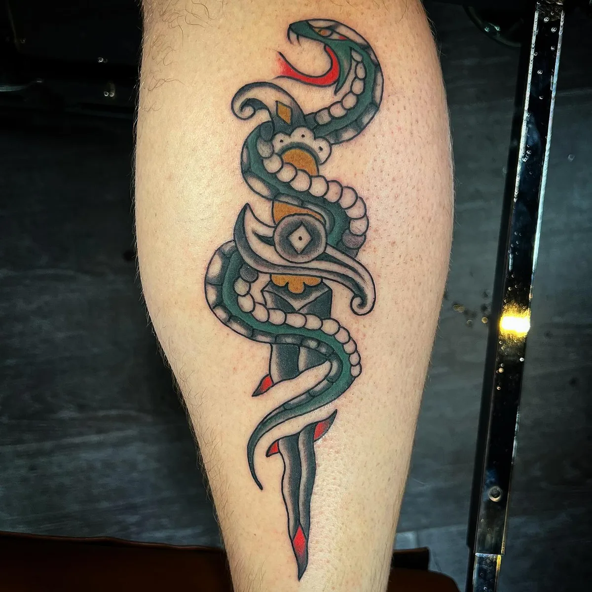 A traditional-style tattoo featuring a snake wrapped around a dagger, rendered in vibrant colours with heavy black outlines and classic shading.
