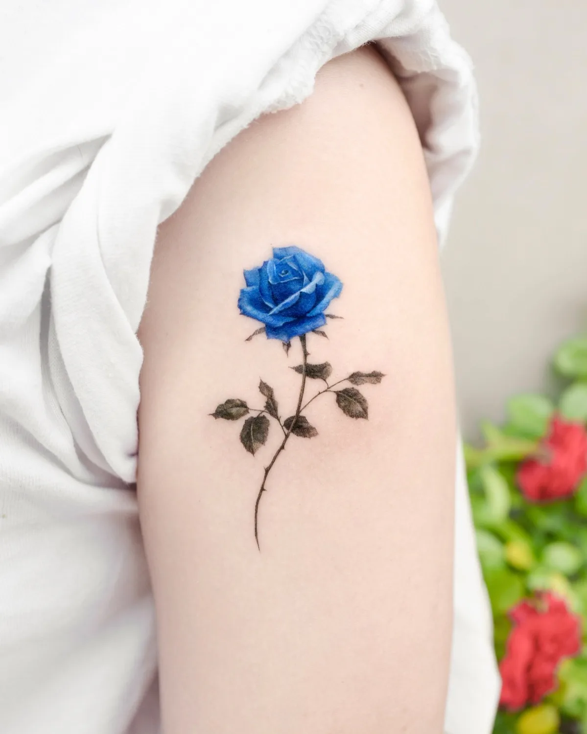 Detailliertes blaues Rosen-Tattoo mit fotorealistischer Schattierung und feiner Linienführung am Oberarm, das zarte Blütenblätter und dimensionale Tiefe zeigt.