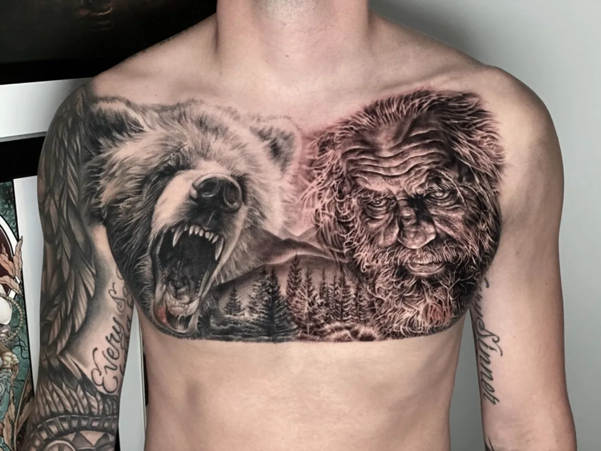 A detailed black and grey tattoo featuring a snarling bear and a portrait of a man with a forest scene in the background.