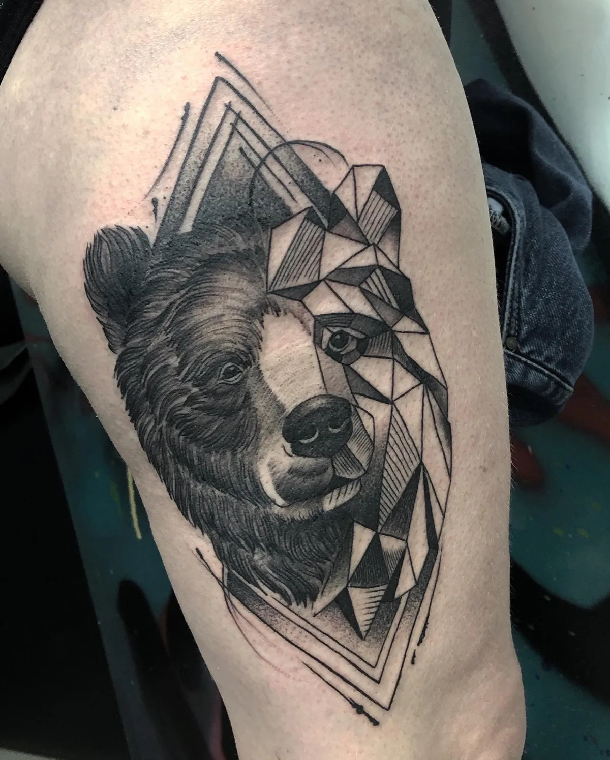 A black and grey tattoo of a bear's face with one half rendered realistically and the other in a geometric style.