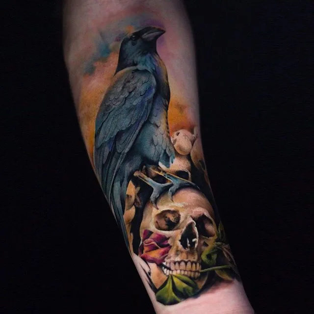 A realistic tattoo showing a crow perched on top of a human skull while holding a rose in its beak, rendered in black and grey.