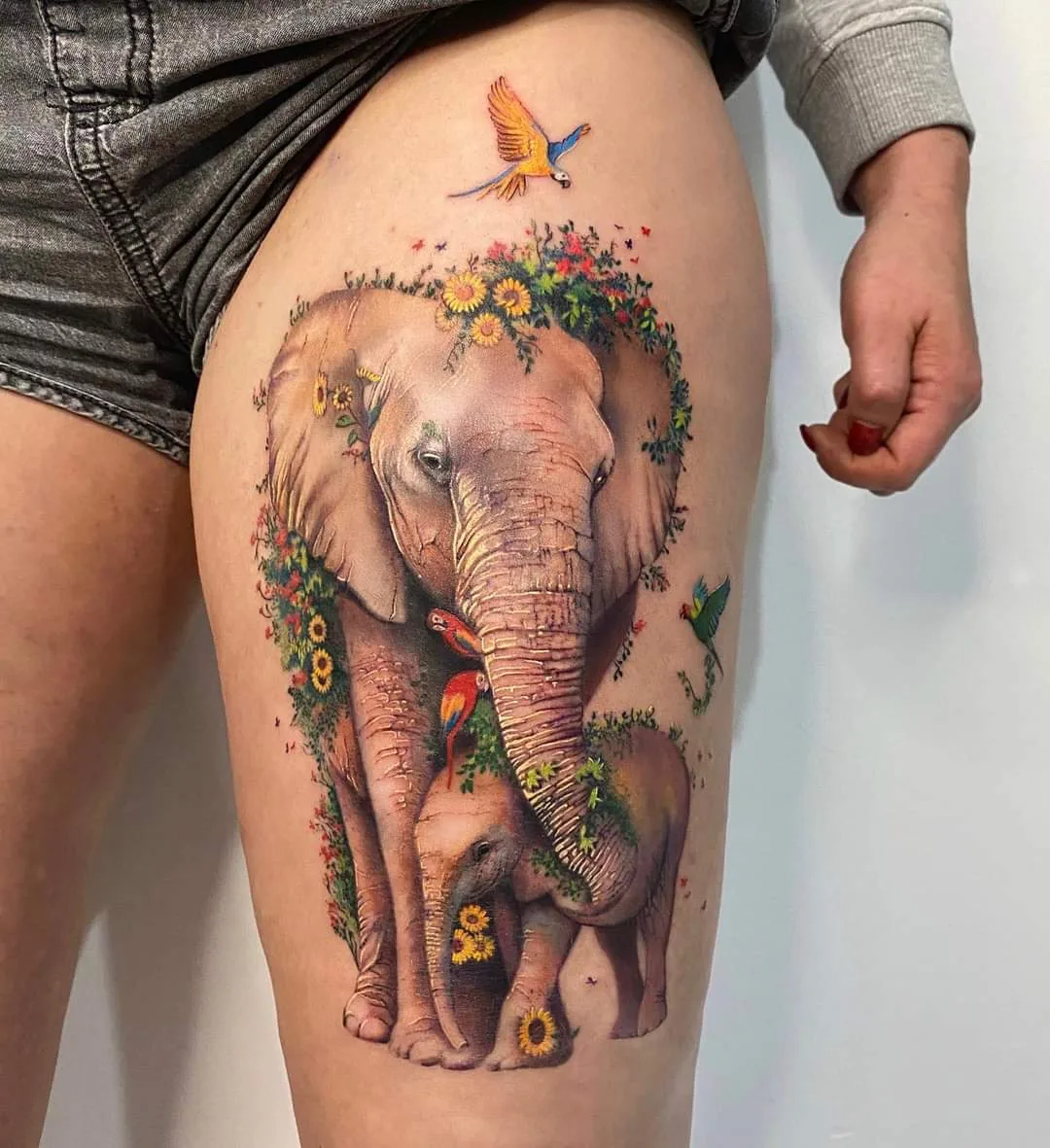 A realistic tattoo of an adult elephant and a calf surrounded by flowers and birds, rendered in black and grey with fine detail.