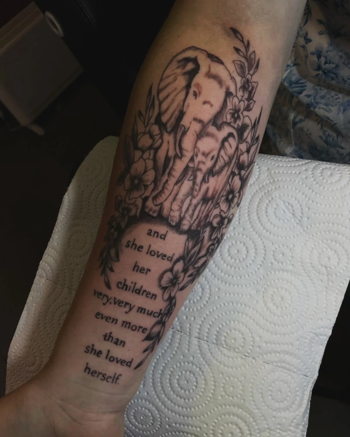 A black and grey forearm tattoo featuring two elephants surrounded by floral elements and an inscribed quote, composed in a soft illustrative style.