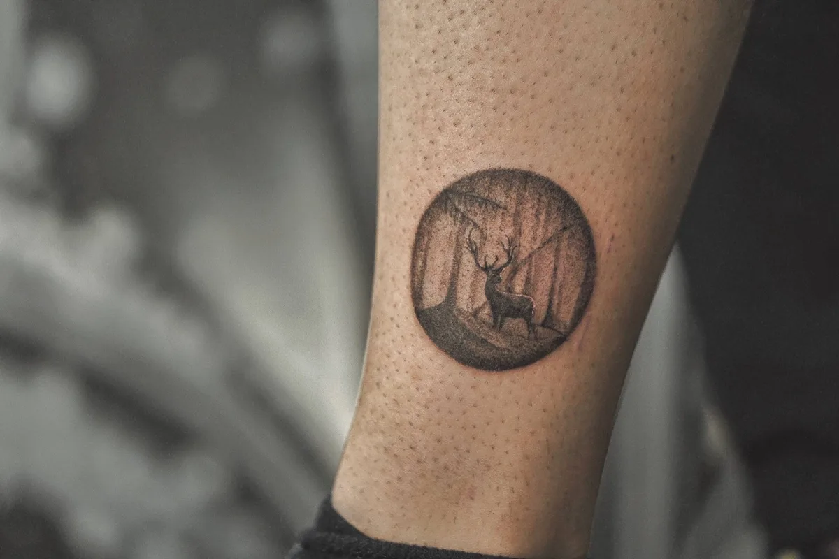A detailed black and grey tattoo of a deer standing in a forest, contained within a circular frame, rendered in a sketch-like style.