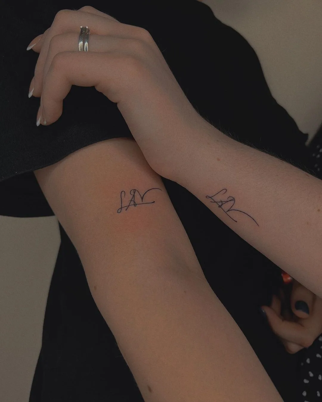 Two forearms each bearing a minimalist tattoo of a personal initial. The designs are matching in style and placement, rendered in fine black ink.