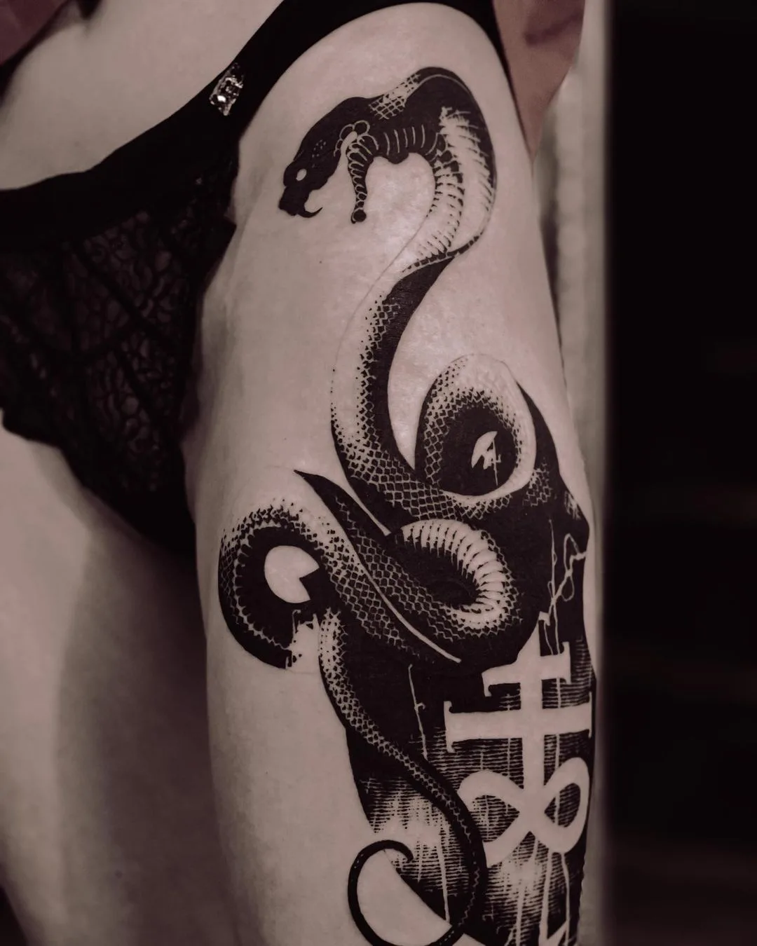A blackwork tattoo in an engraving style showing a detailed snake coiled around a cross, the linework dense and precise.
