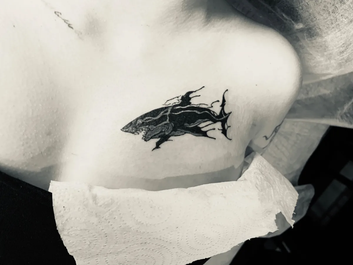 A blackwork sketch-style tattoo of a shark placed along the collarbone, rendered with loose expressive linework.