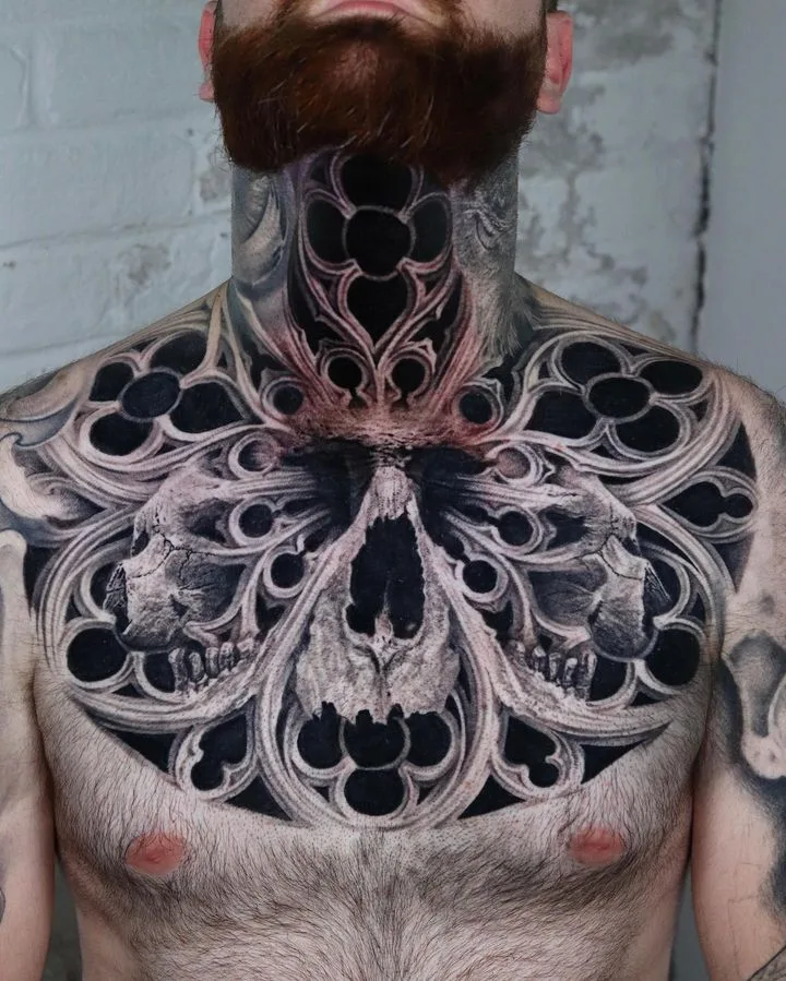 A large, intricate blackwork tattoo featuring a skull and gothic ornamental design covering the chest and extending up the neck.