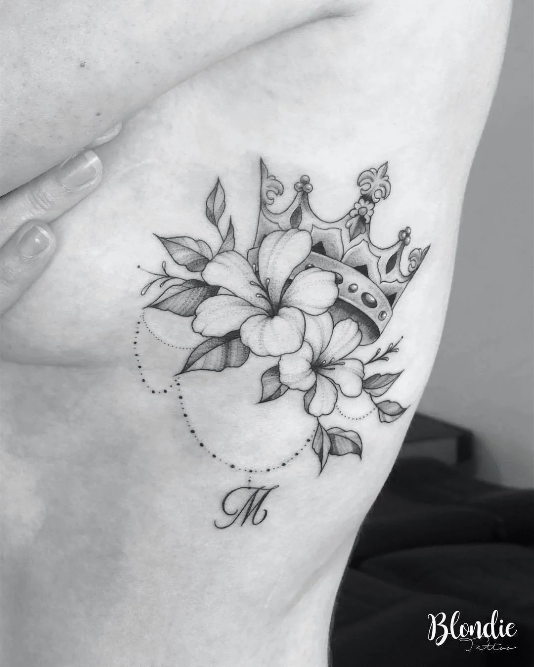 Tatuaggio in bianco e nero sul costato con una corona, fiori e una lettera M in corsivo, composizione delicata e femminile.