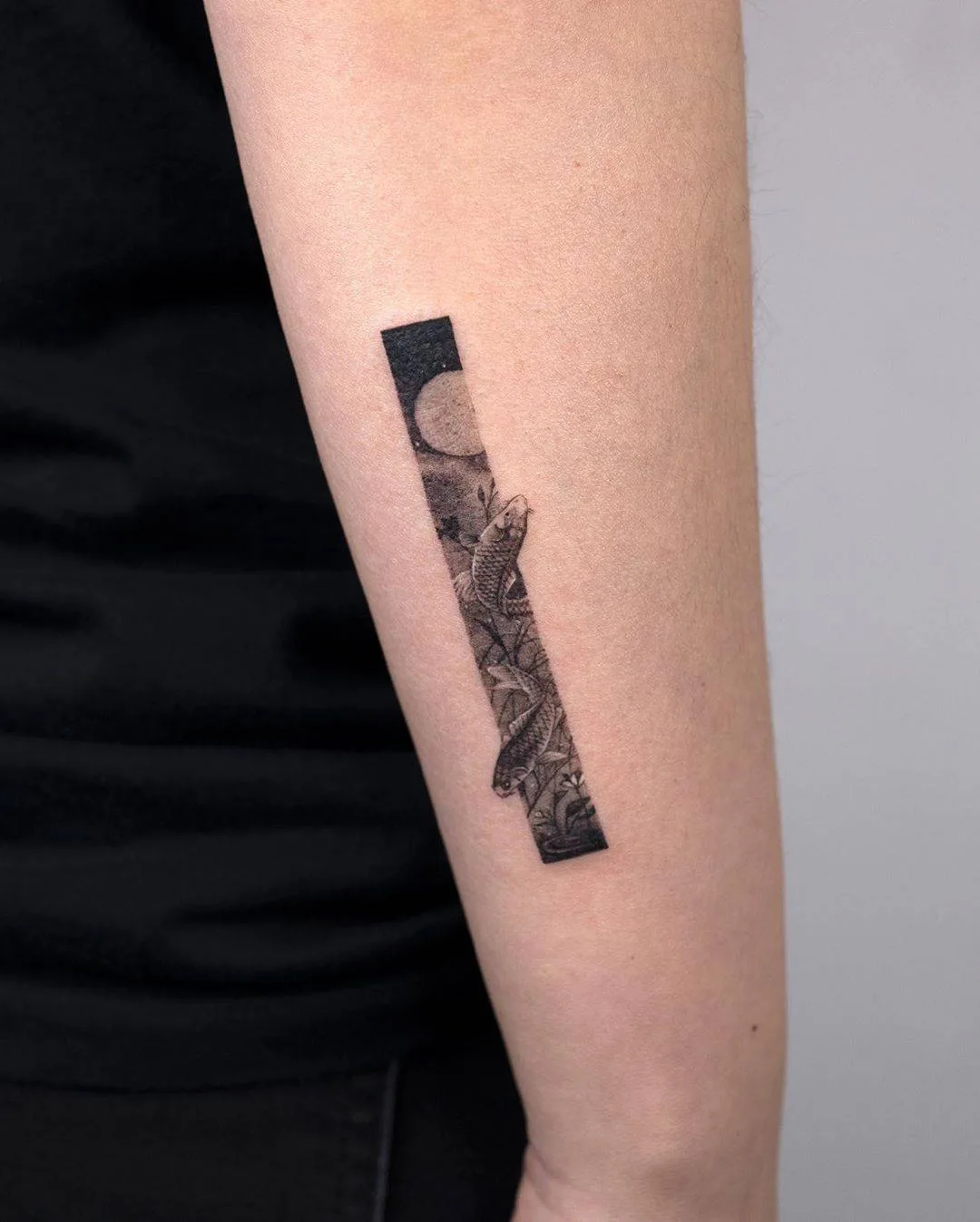 A black and grey tattoo of a fish swimming in a rectangular frame with a moon and nature elements.
