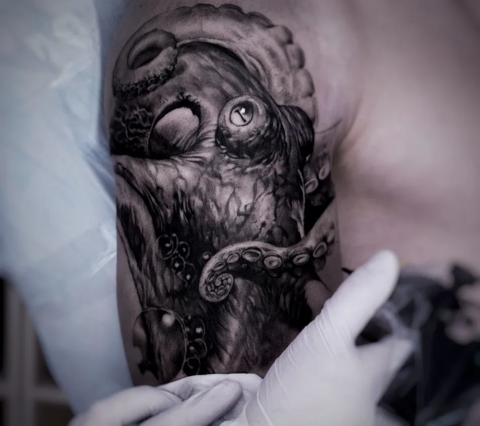 A black and grey octopus tattoo being applied to the upper arm, with a gloved hand holding the tattoo machine visible in the frame.
