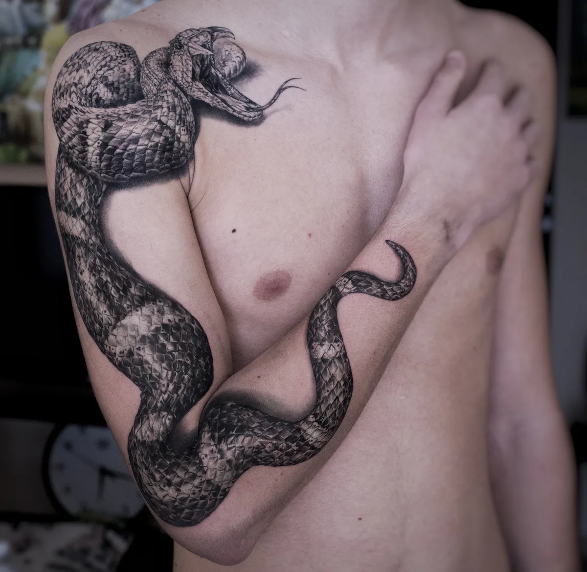 A realistic black and grey snake tattoo that wraps continuously around the arm and shoulder, with detailed scale shading and a naturalistic pose.