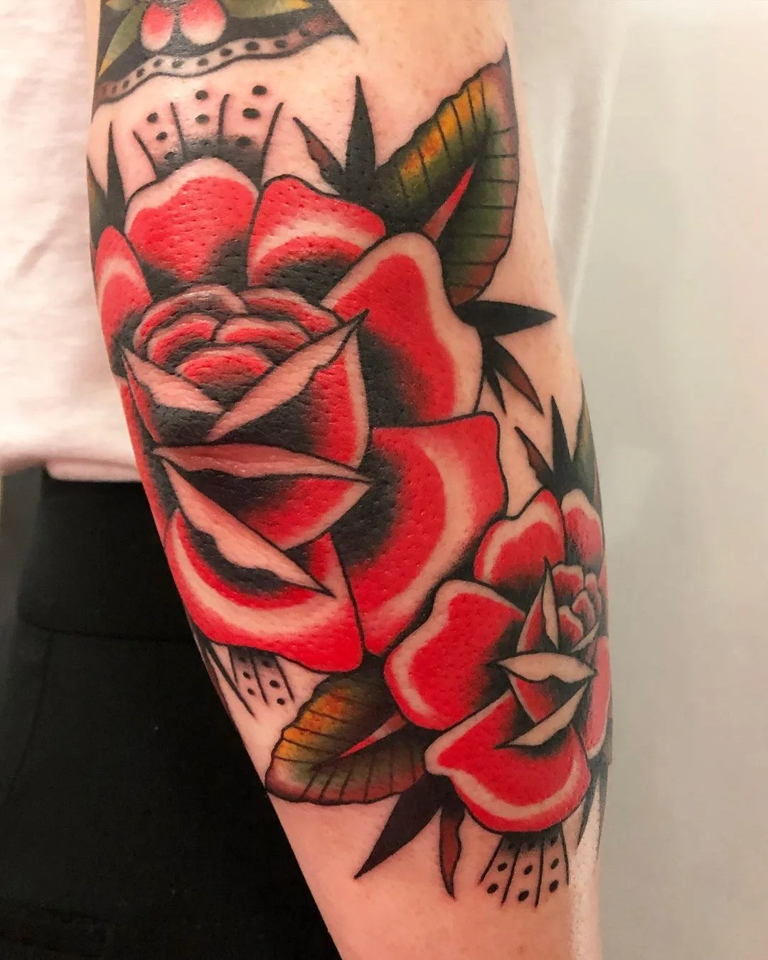 Zwei leuchtend rote Rosen mit schwarzen Schattierungen und grünen Blättern im klassischen traditionellen Tattoo-Stil auf der Haut.