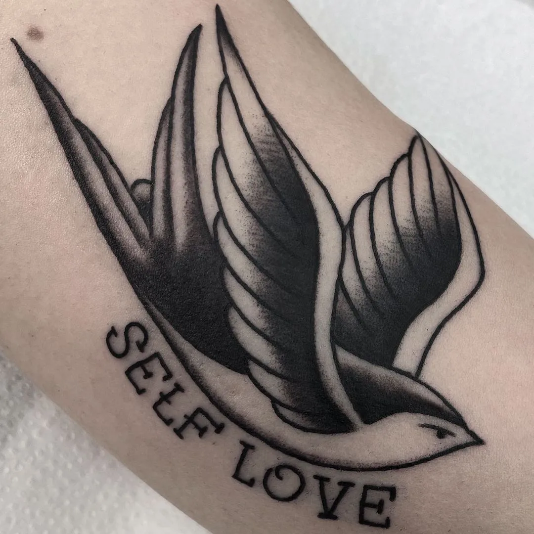 Ein Traditional-Tattoo einer Schwalbe in Schwarz-Grau mit dem Schriftzug 'Self Love' darunter.