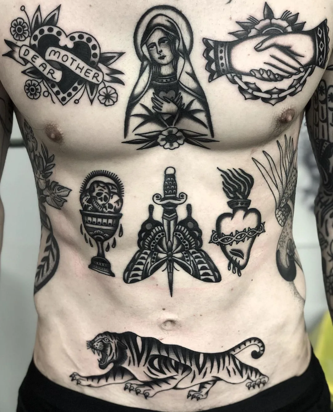 A collection of traditional style tattoos covering the chest and abdomen, including a tiger, dagger, and religious imagery in bold classic colours.