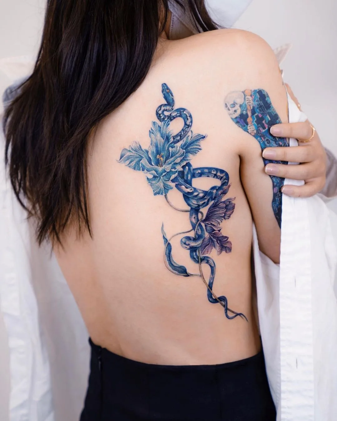 A delicate illustrative fine line tattoo of a blue snake intertwined with a flower, with light shading and minimal linework.