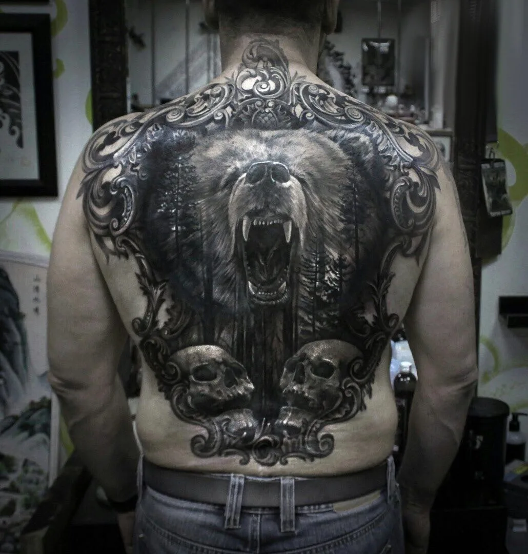A detailed black and grey tattoo of a roaring bear in a forest scene framed by ornate scrollwork and skulls.