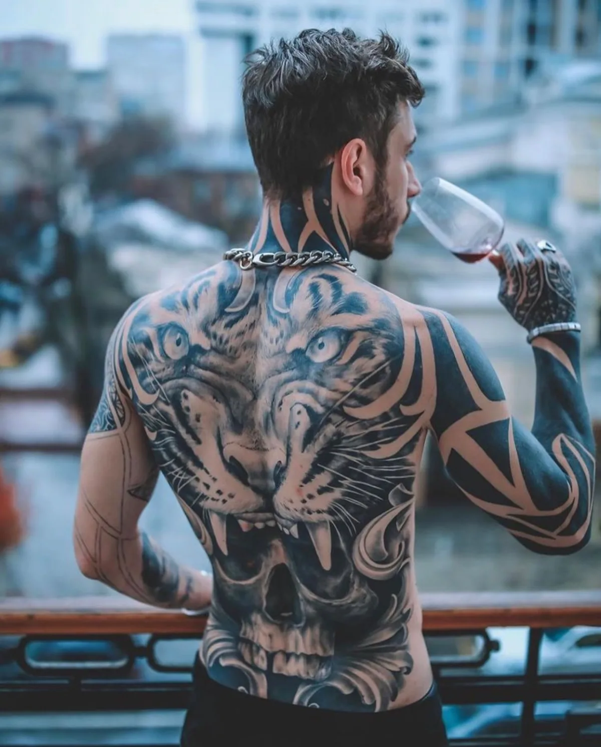 A large black and grey tattoo covering the man's back, featuring a tiger, skull, and geometric patterns, with blackout work extending to the neck and arms.