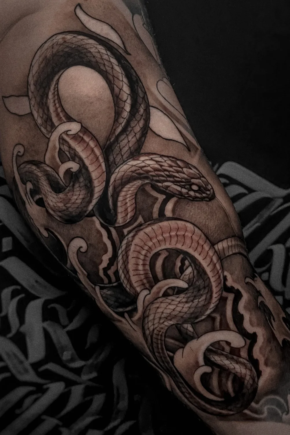 A detailed black and grey tattoo of a snake coiled around itself with flowing water elements, evoking themes of renewal and cyclical energy.