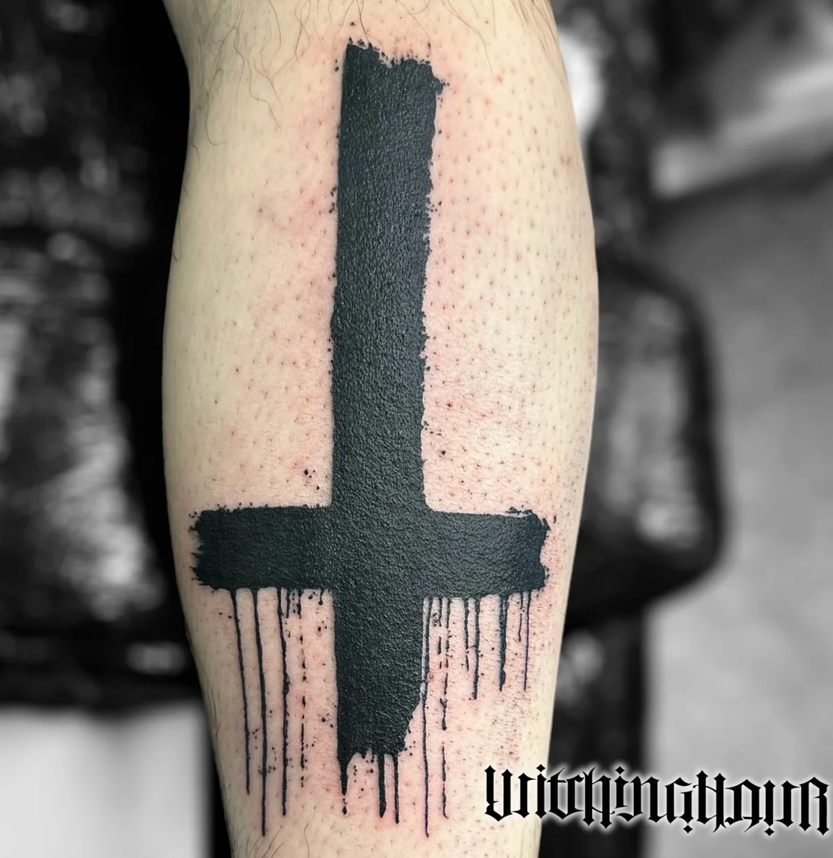 A large, inverted cross tattoo in black ink with a dripping effect. The tattoo has a rough, textured appearance.
