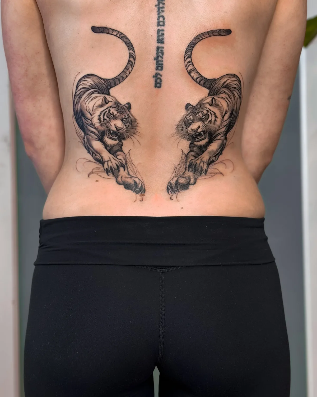 A tattoo featuring two realistic tigers facing each other on the lower back, with Hebrew lettering running down the spine.