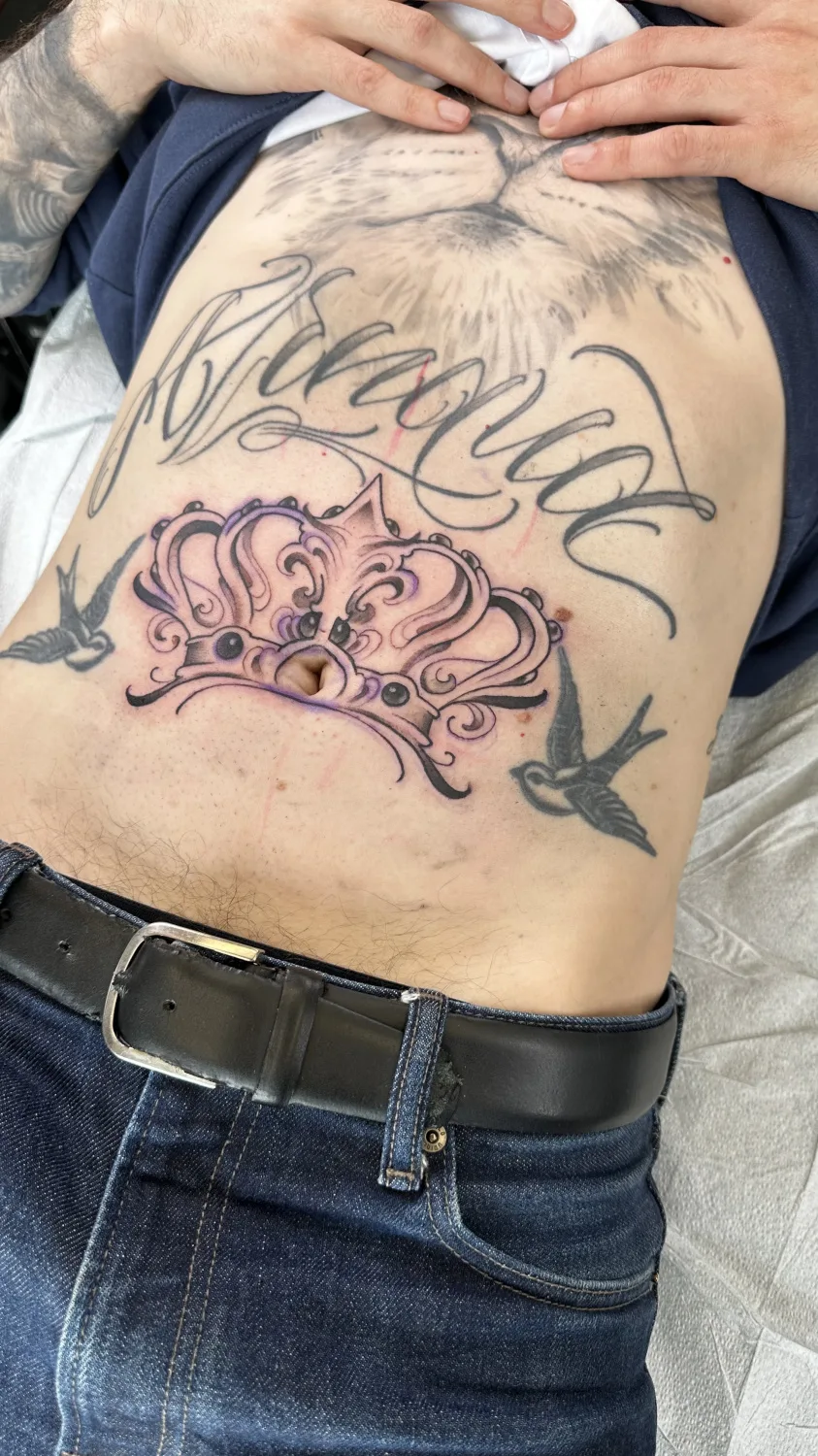 Ein Schwarz-Grau-Tattoo mit einer Krone, Schwalben und Schriftzug auf dem Bauch.