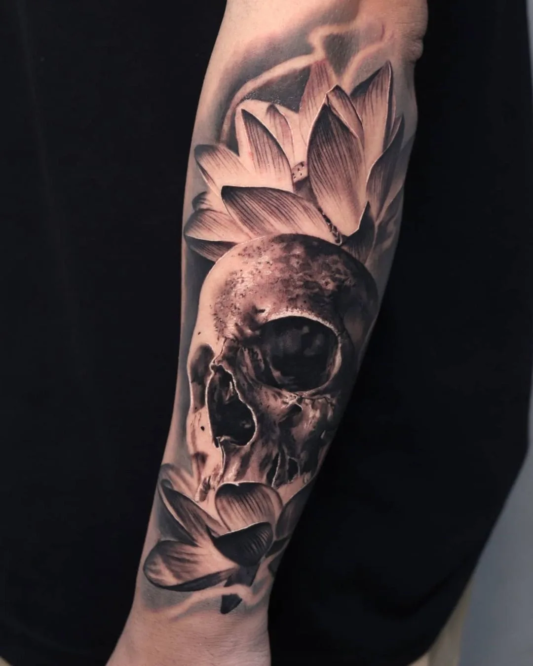 A detailed black and grey tattoo featuring a realistic skull surrounded by blooming lotus flowers.