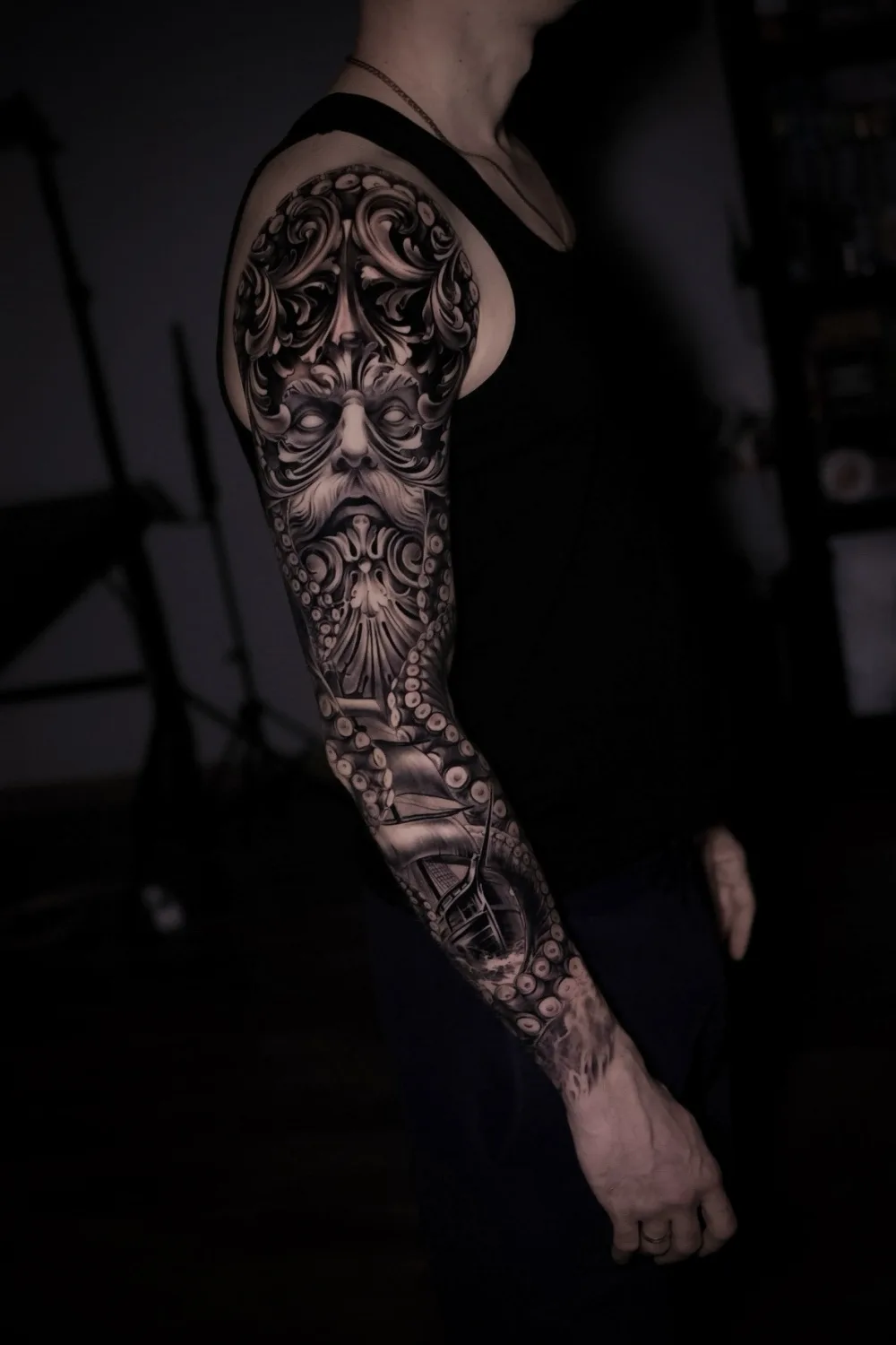 A detailed black and grey full sleeve tattoo featuring an ornate face, a sailing ship, and octopus tentacles wrapping around the arm.