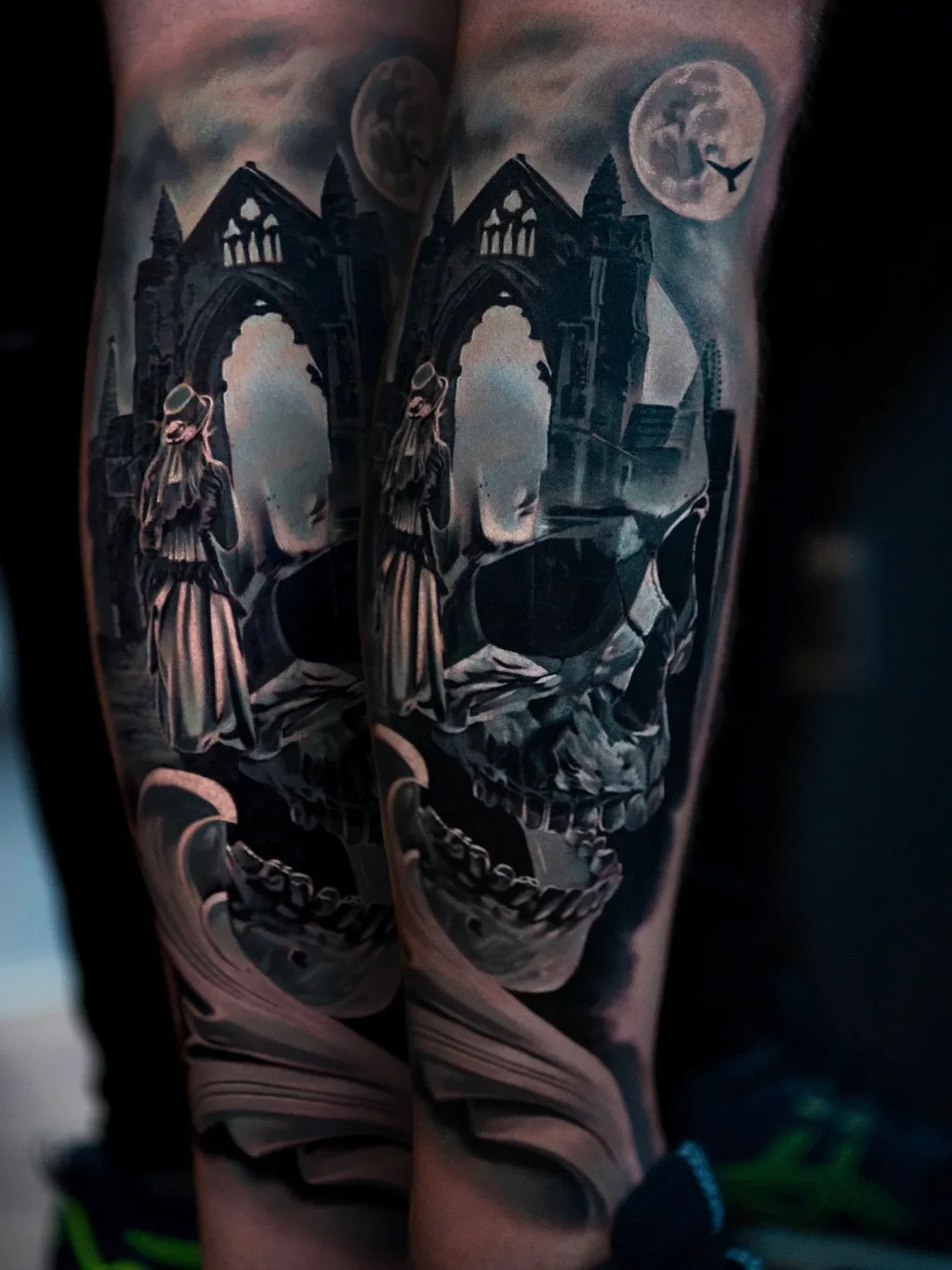 A detailed black and grey tattoo featuring a skull, gothic architectural elements, and a moonlit sky in the background.