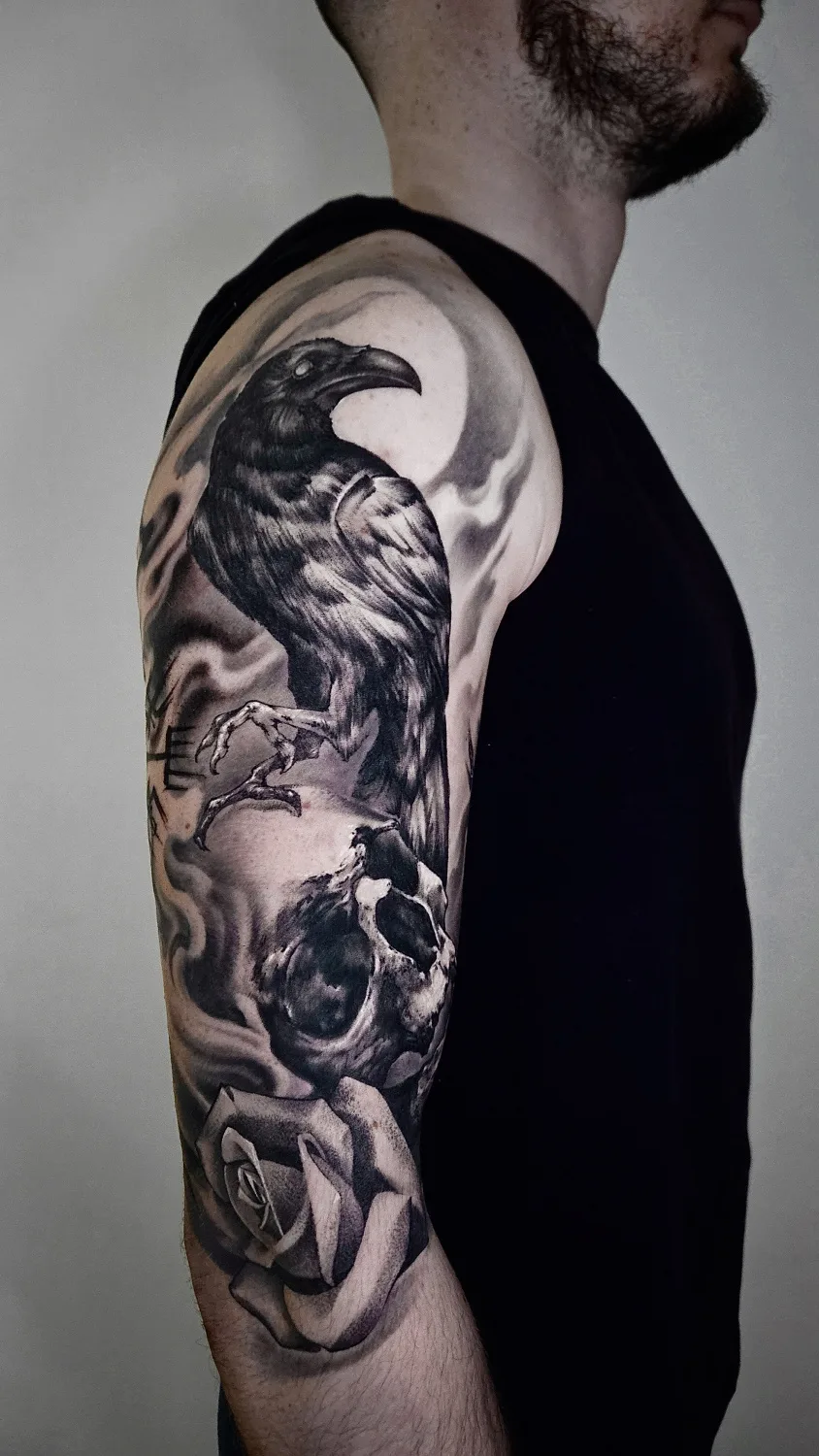 A black and grey tattoo showing a raven perched above a skull and a rose, with a smoky atmospheric background, rendered in a realistic style.