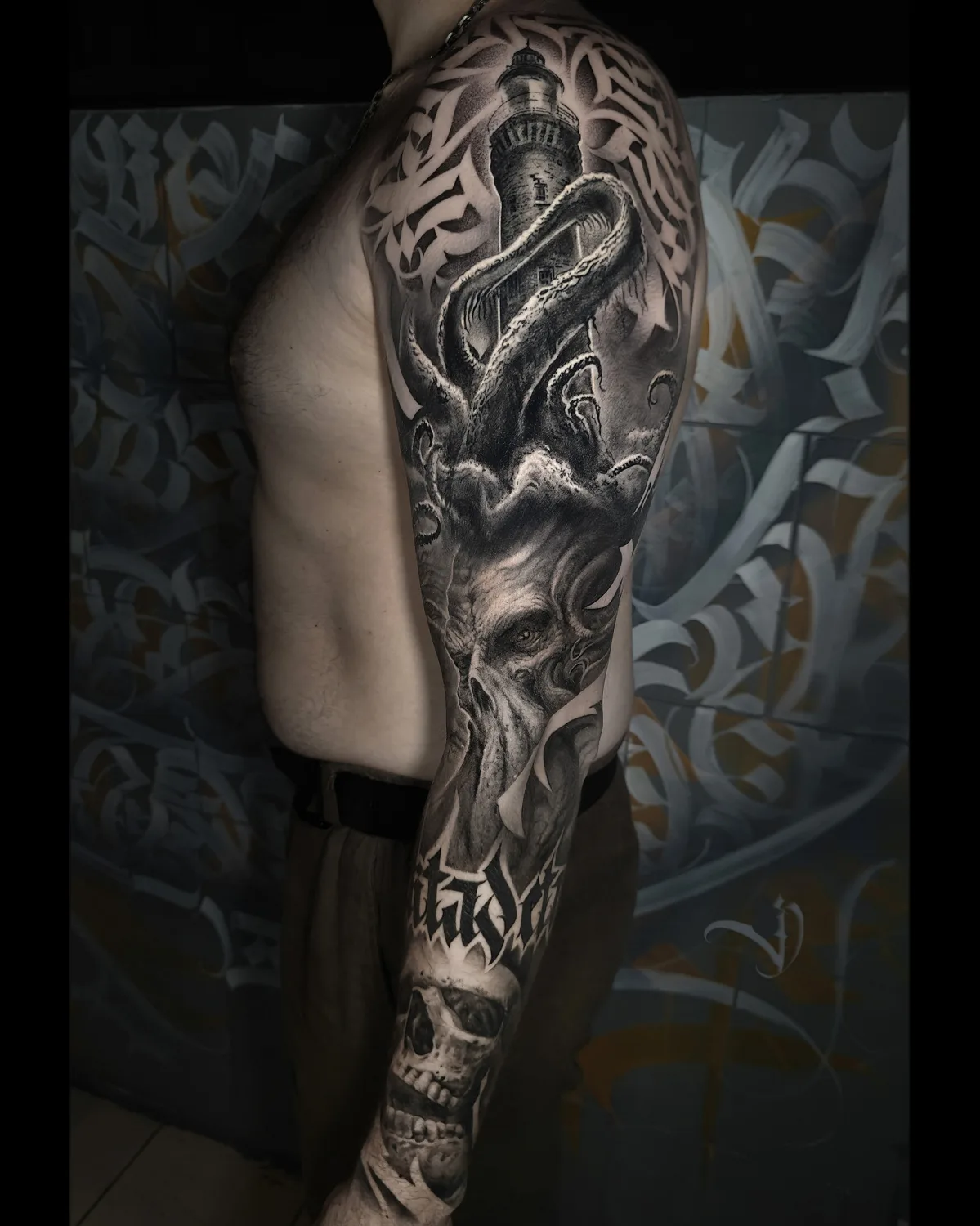 A detailed black and grey full sleeve tattoo depicting a lighthouse being attacked by a giant octopus, with a skull at the bottom and calligraphy elements.