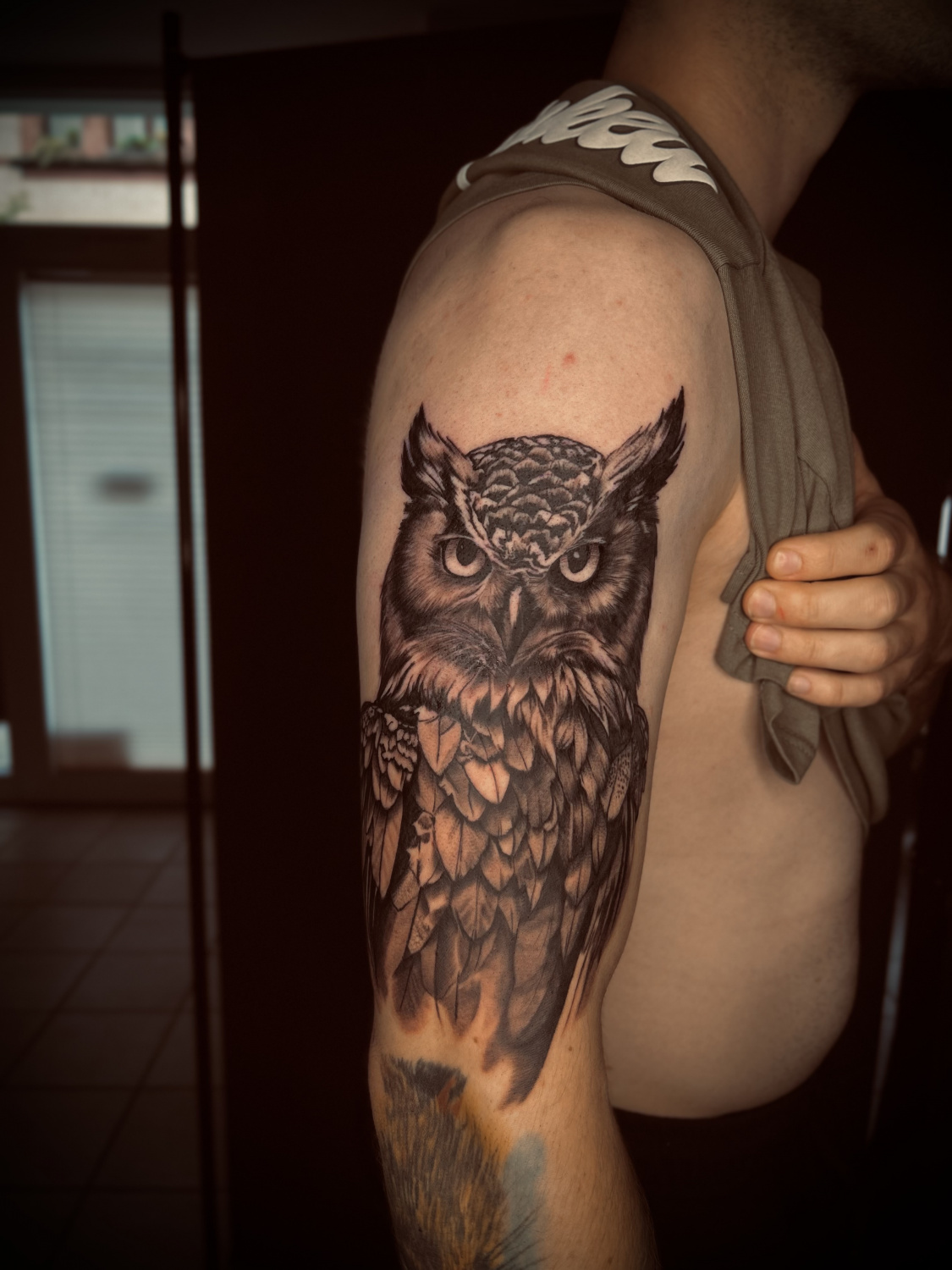 A detailed black and grey owl tattoo on the upper arm, showcasing realistic shading and intricate layered feather details.