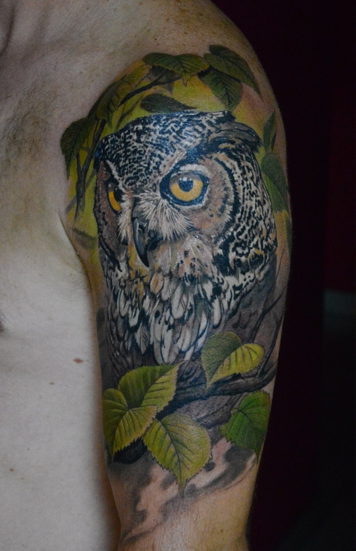 A realistic owl tattoo surrounded by autumn leaves, featuring detailed shading and warm color tones rendered in a painterly style.