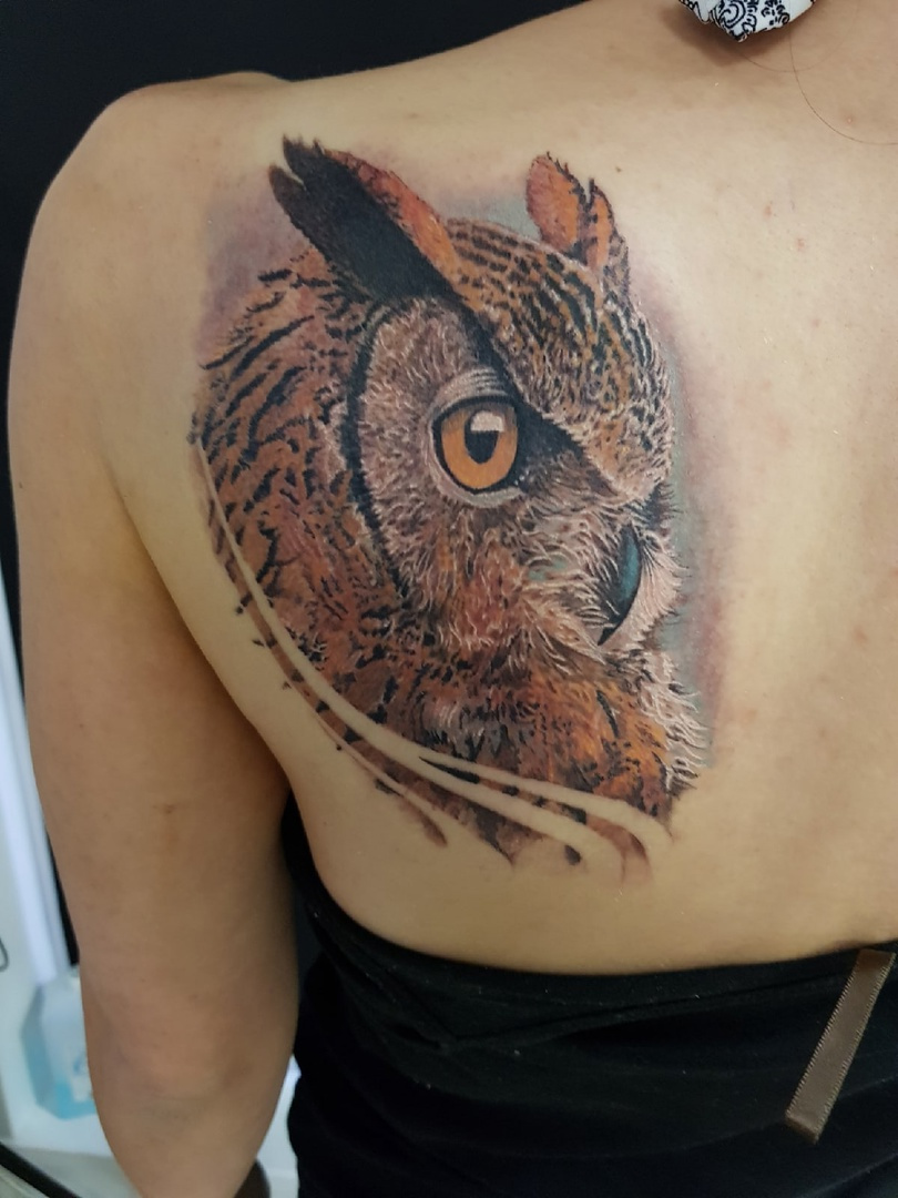 A realistic tattoo of an owl's head showcasing detailed layered feather textures and vibrant warm colors on a large area of skin.