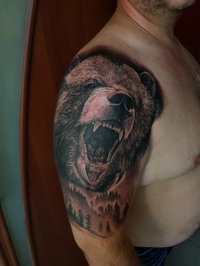 A detailed black and grey tattoo of a roaring bear's head with a forest scene below.