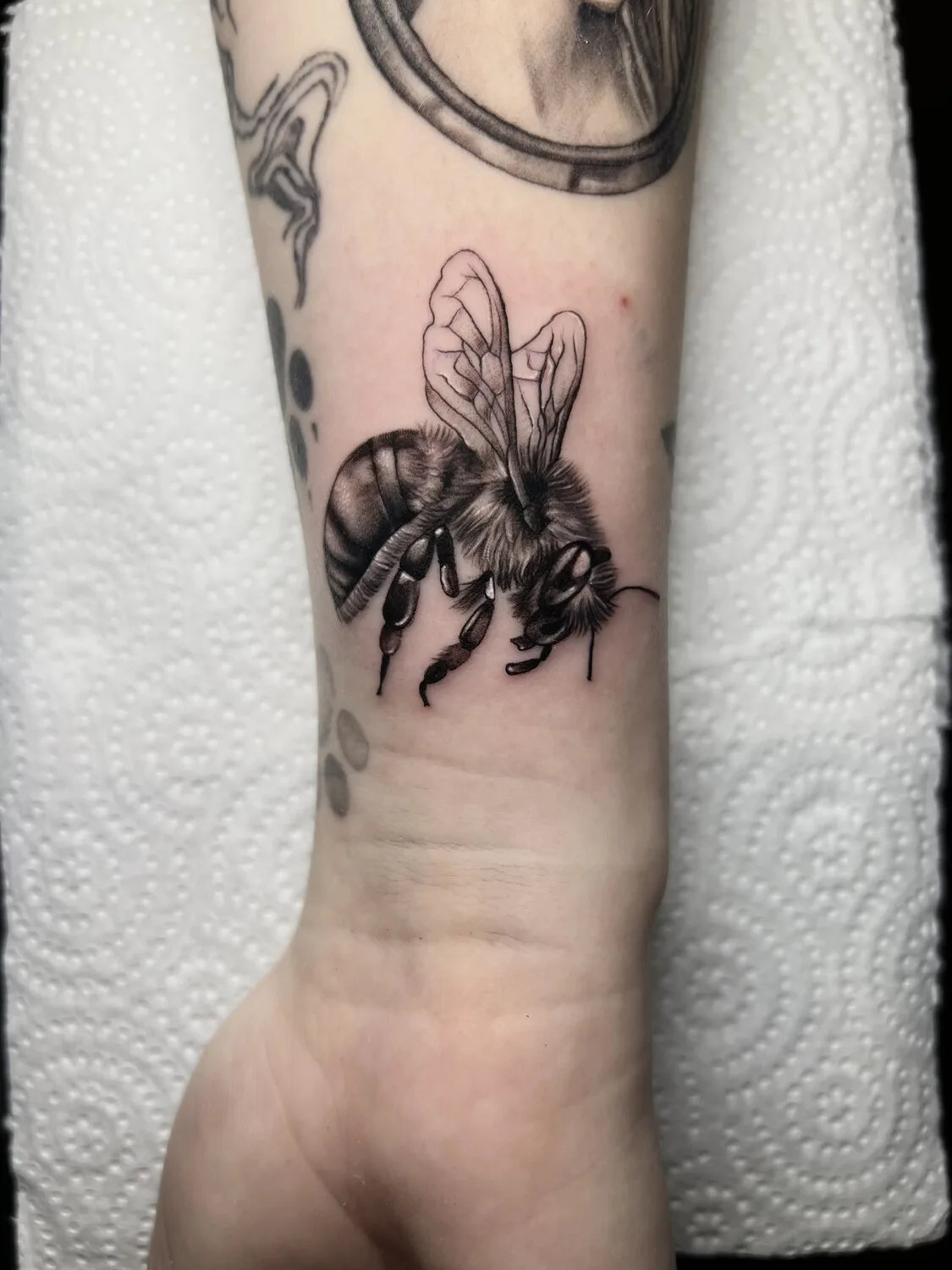 A detailed black and grey realistic bee tattoo placed on the wrist, with careful shading on the thorax and abdomen and fine linework on the wings.