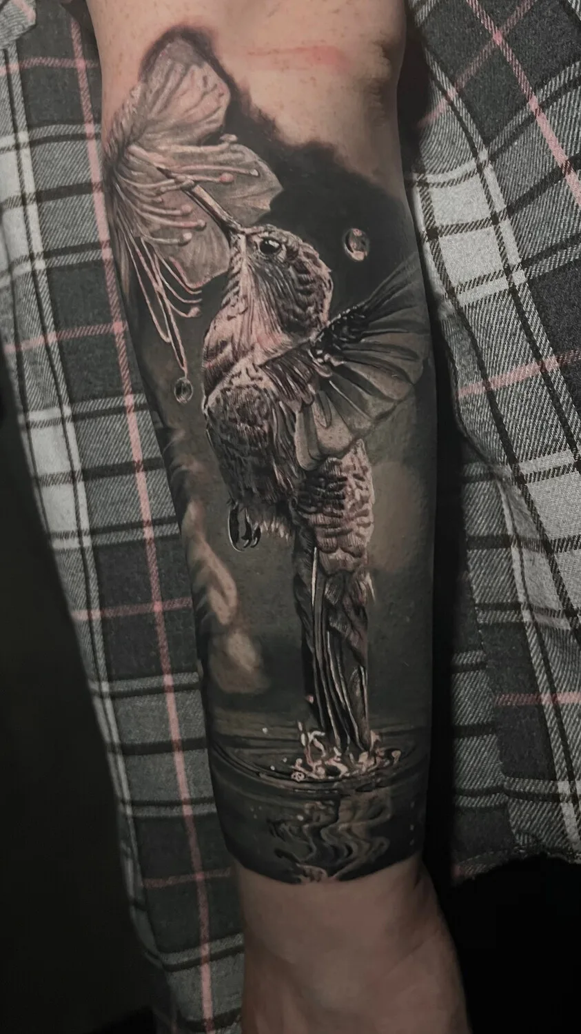A black and grey realistic tattoo of a hummingbird drinking from a flower, with finely rendered water droplets and reflective highlights suggesting stillness and memory.