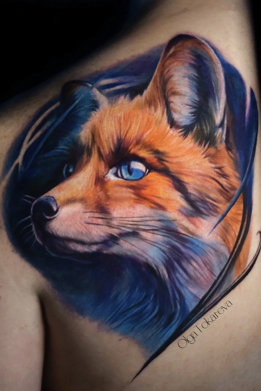 A close-up of a watercolor-style fox head tattoo with realistic fur texture and bright blue eyes, surrounded by soft colour washes.