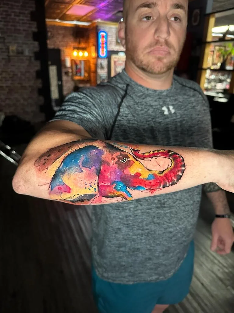 A colourful watercolour-style tattoo of an elephant's head and trunk, using loose painterly brushwork and vibrant blues, purples and pinks.