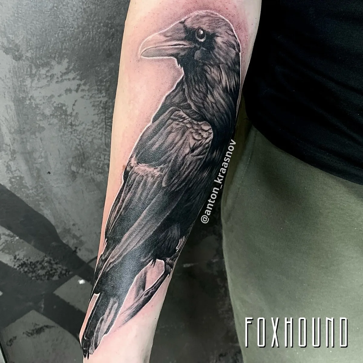A black and grey realistic tattoo of a raven perched on a bare branch, with intricate feather detail and subtle shading that captures the iridescent quality of the plumage.