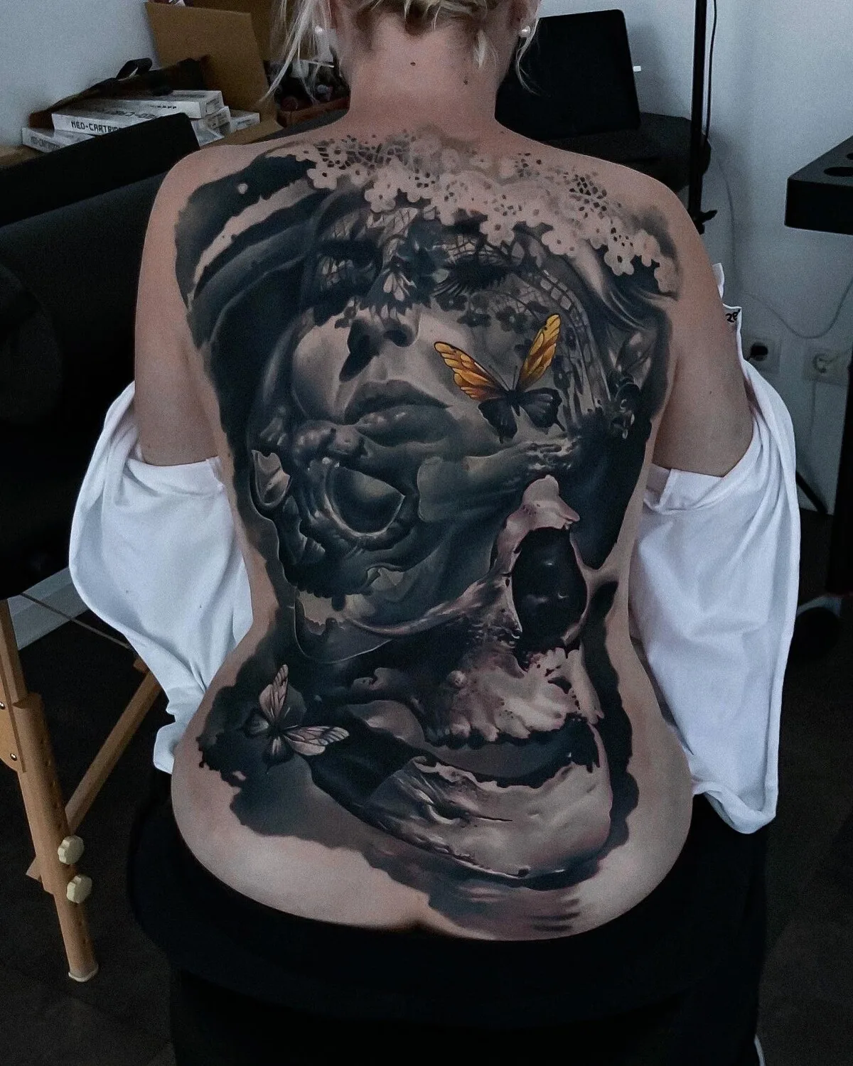A large, detailed black and grey tattoo featuring a skull, a human face, and butterflies, highly realistic with intricate shading.