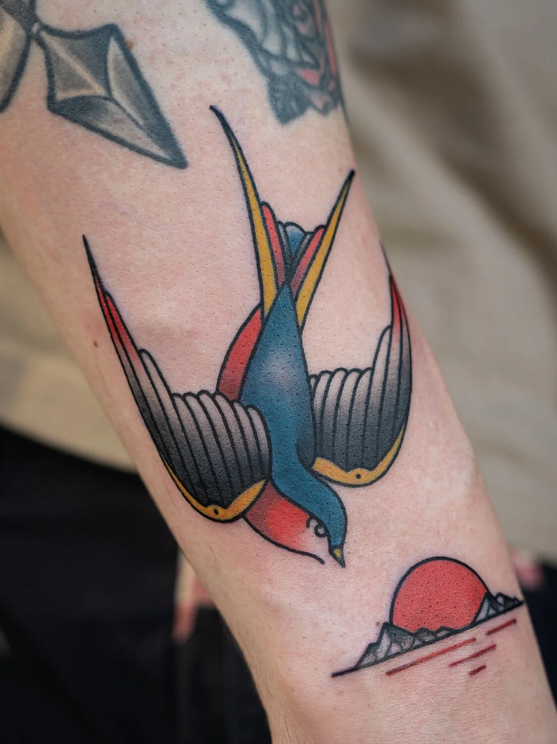 Ein klassisches Traditional-Tattoo mit einer Schwalbe im Flug vor einem Sonnenuntergang, ausgeführt in kräftigen Linien und satten Farben.