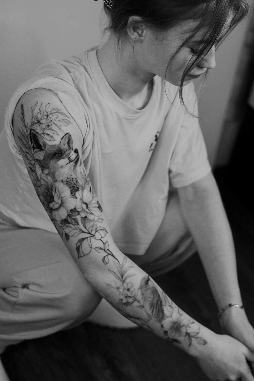 A black and grey tattoo on the forearm featuring a fox surrounded by flowers and a bird, rendered in detailed realistic shading.