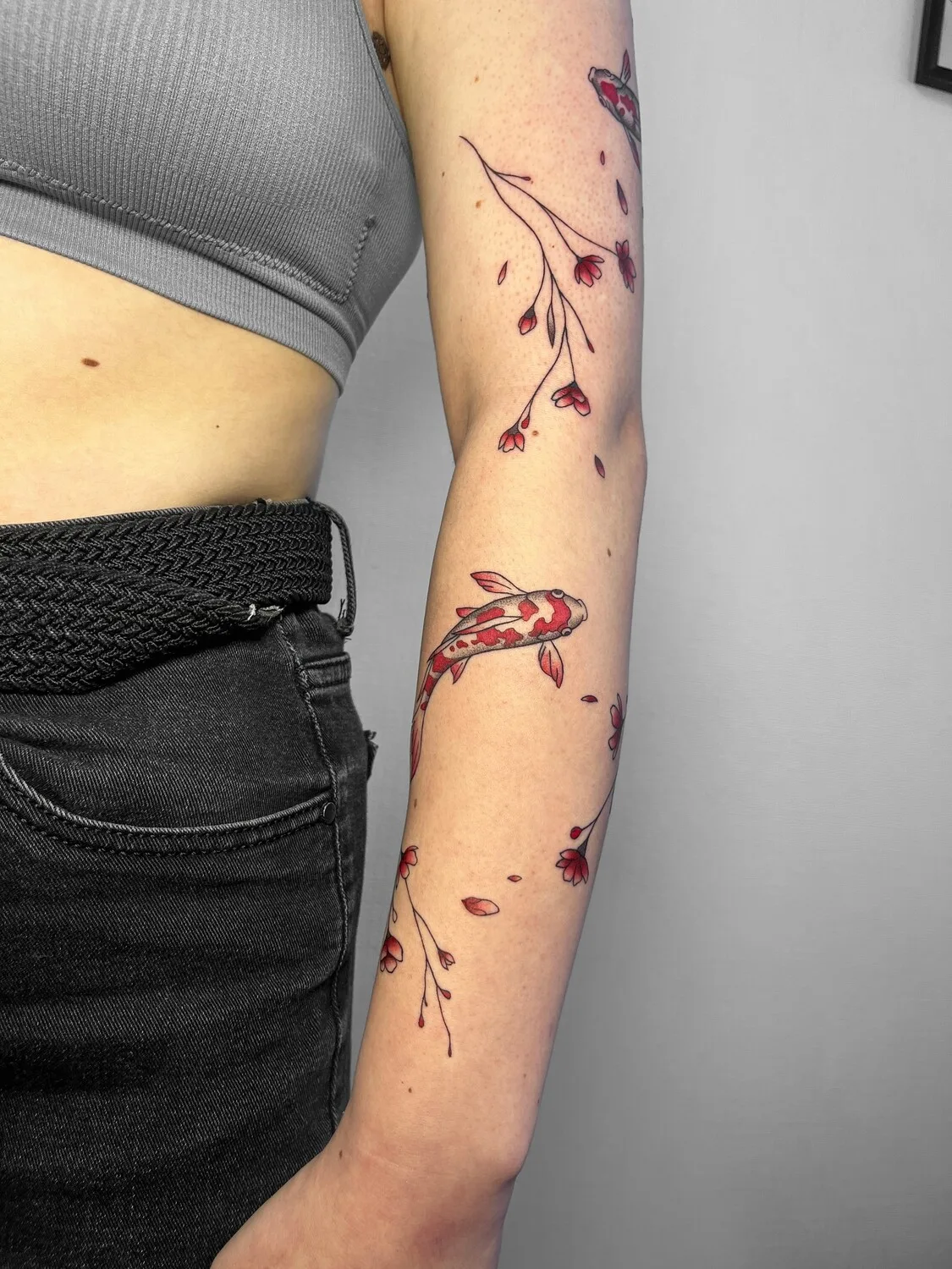 A colourful tattoo on the forearm showing a koi fish swimming among cherry blossom branches in a Japanese-inspired style.