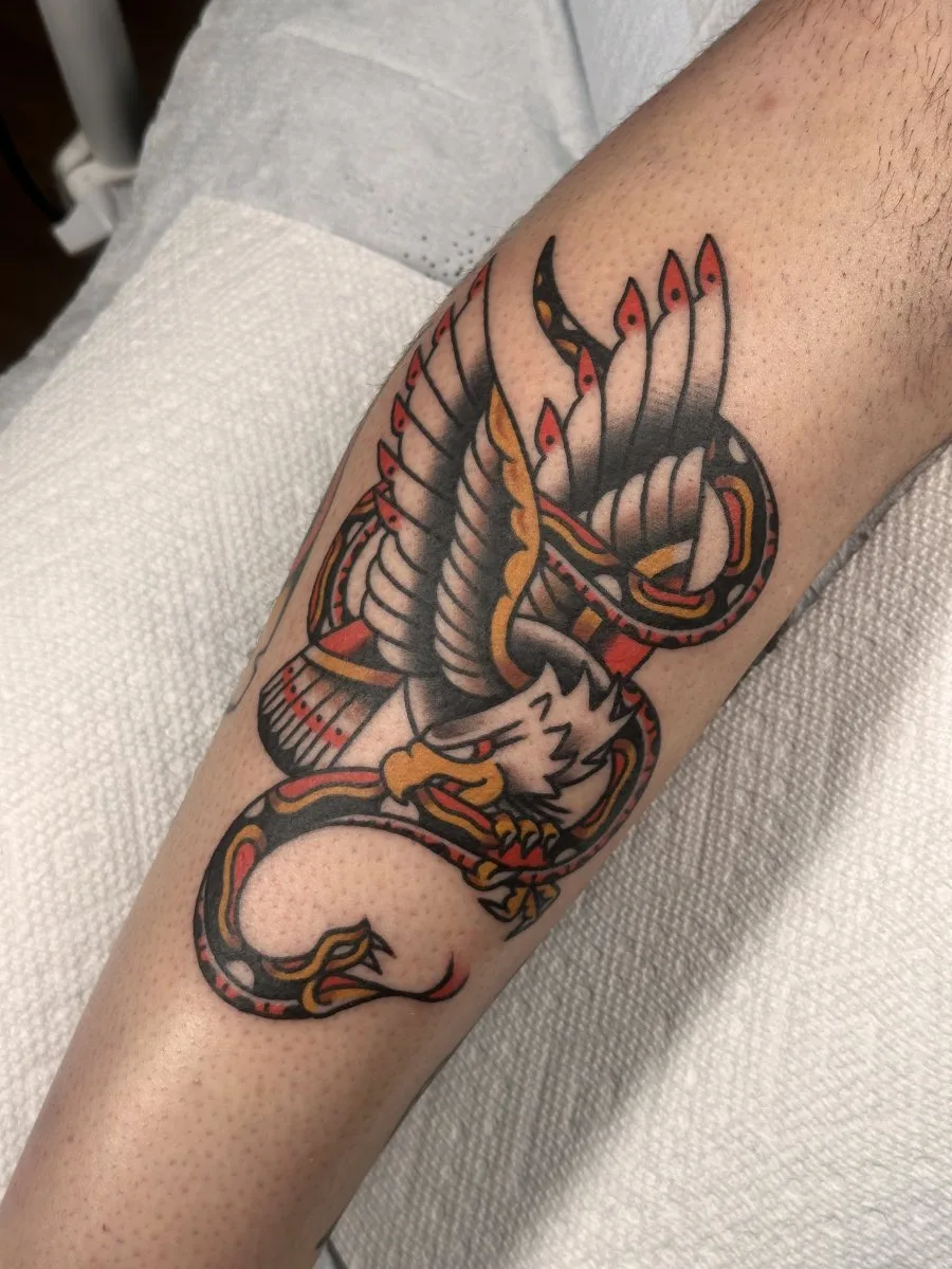 A traditional style tattoo of an eagle with spread wings and talons entwined with a snake wrapping around the leg, with bold black outlines and red, yellow, and grey fills.