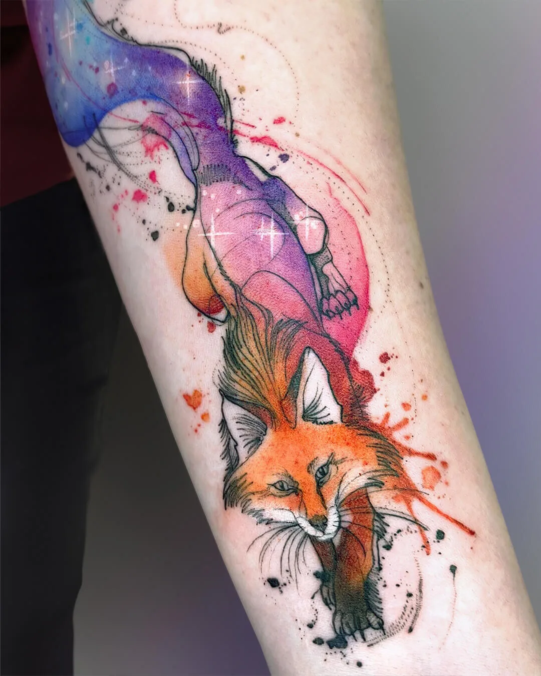 A watercolor tattoo of a fox with a long, colourful cosmic tail, featuring splashes of paint-like colour and fine linework.