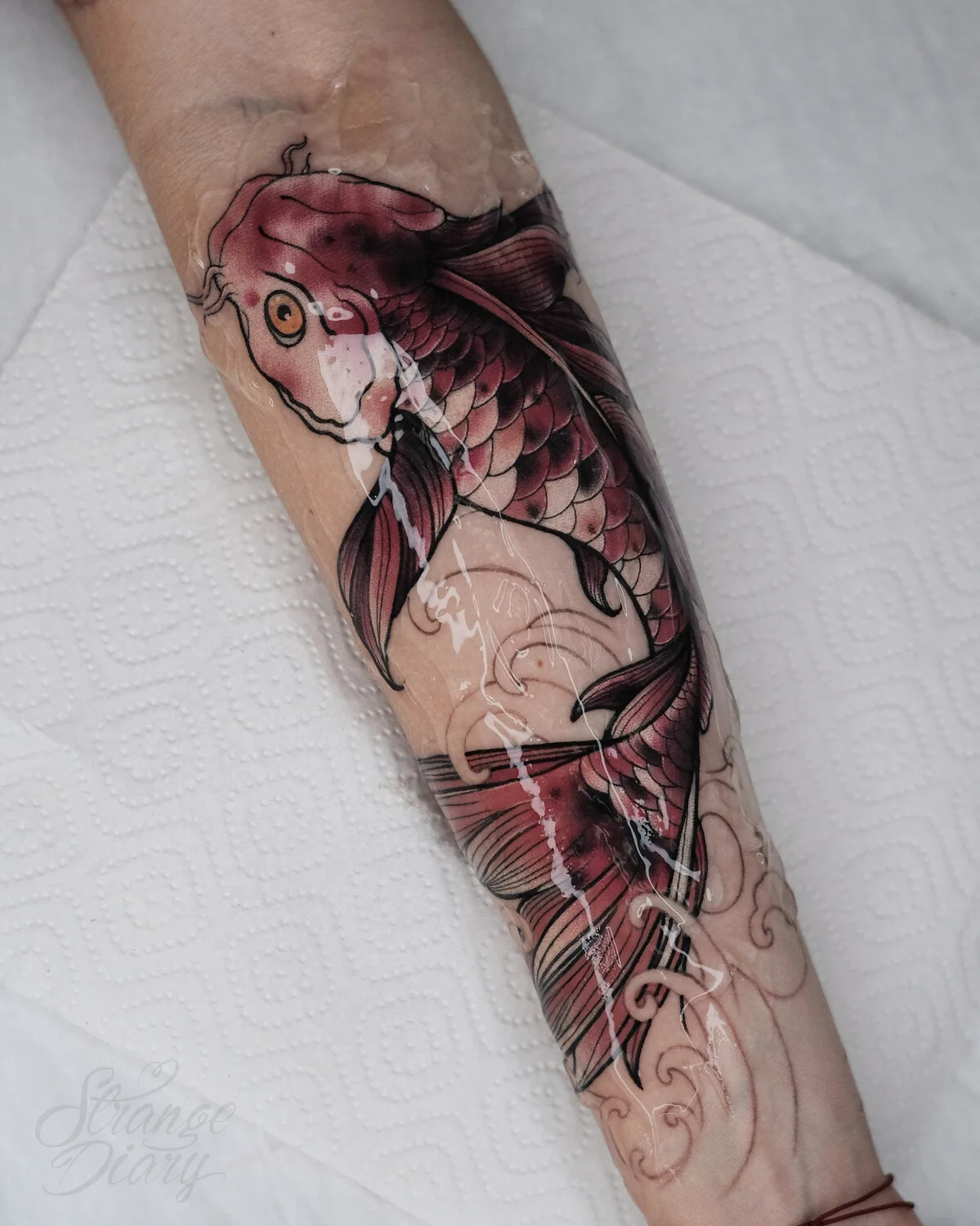 A tattoo on an arm featuring a koi fish with watercolour-style shading, colours bleeding outward into flowing wave shapes.