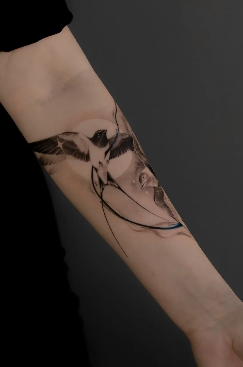 Ein Schwarz-Grau-Unterarm-Tattoo zweier Schwalben im Flug vor einem sanft leuchtenden Mond und Sternen, mit feinem Linienwerk und blauen Akzentstrichen.