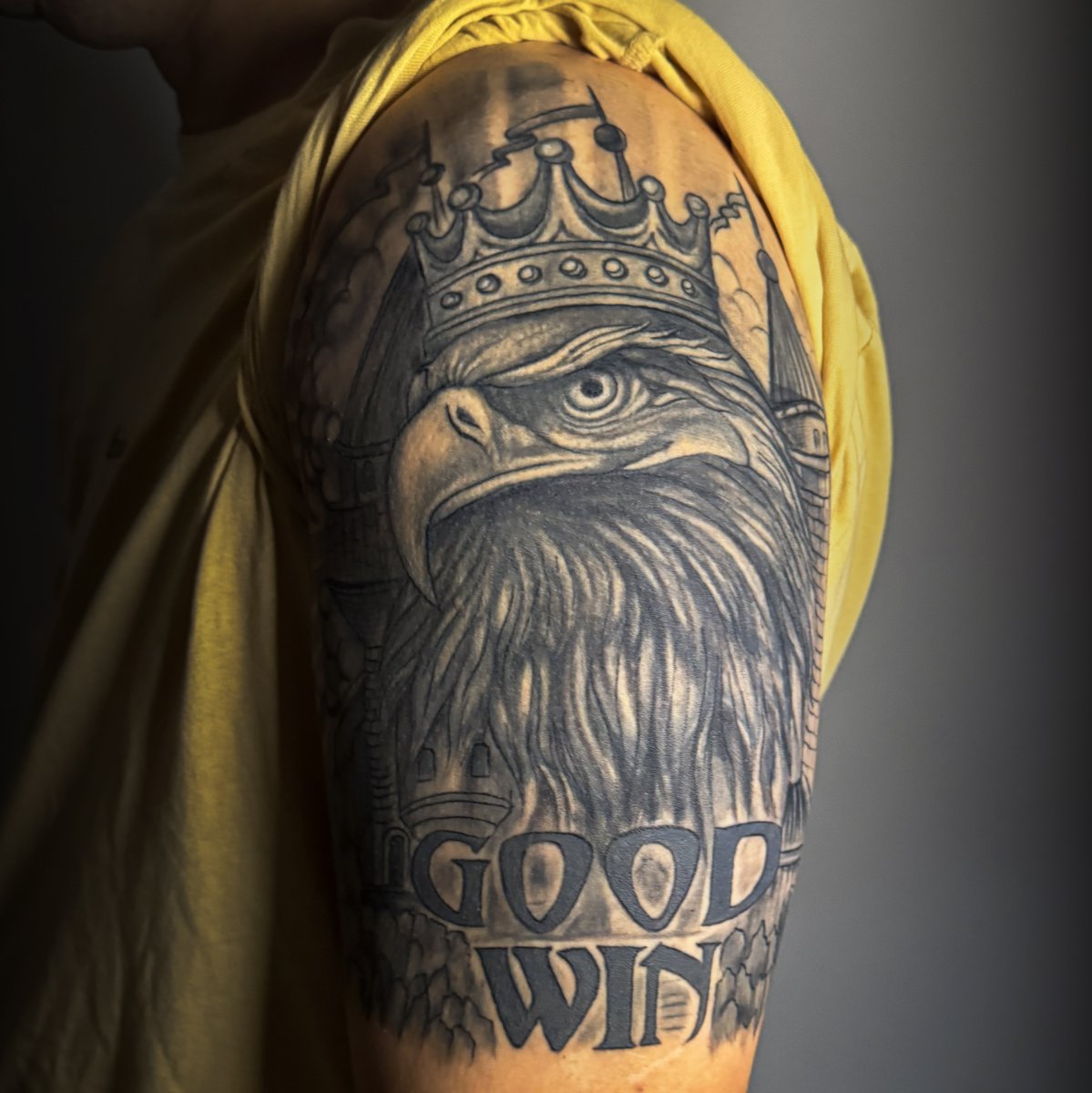 Tatuaggio realistico in bianco e nero di una testa d'aquila con corona, silhouette di castello sullo sfondo e scritta 'GOOD WIN' sul braccio superiore.
