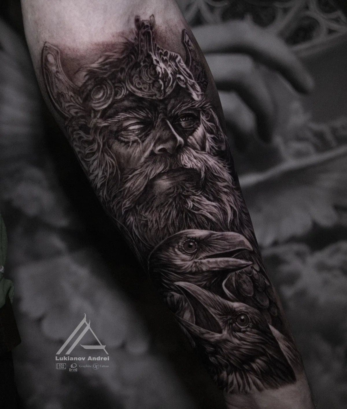 A black and grey tattoo depicting a bearded Norse god figure flanked by two ravens, rendered in a realistic illustrative style with fine detail in the feathers and face.
